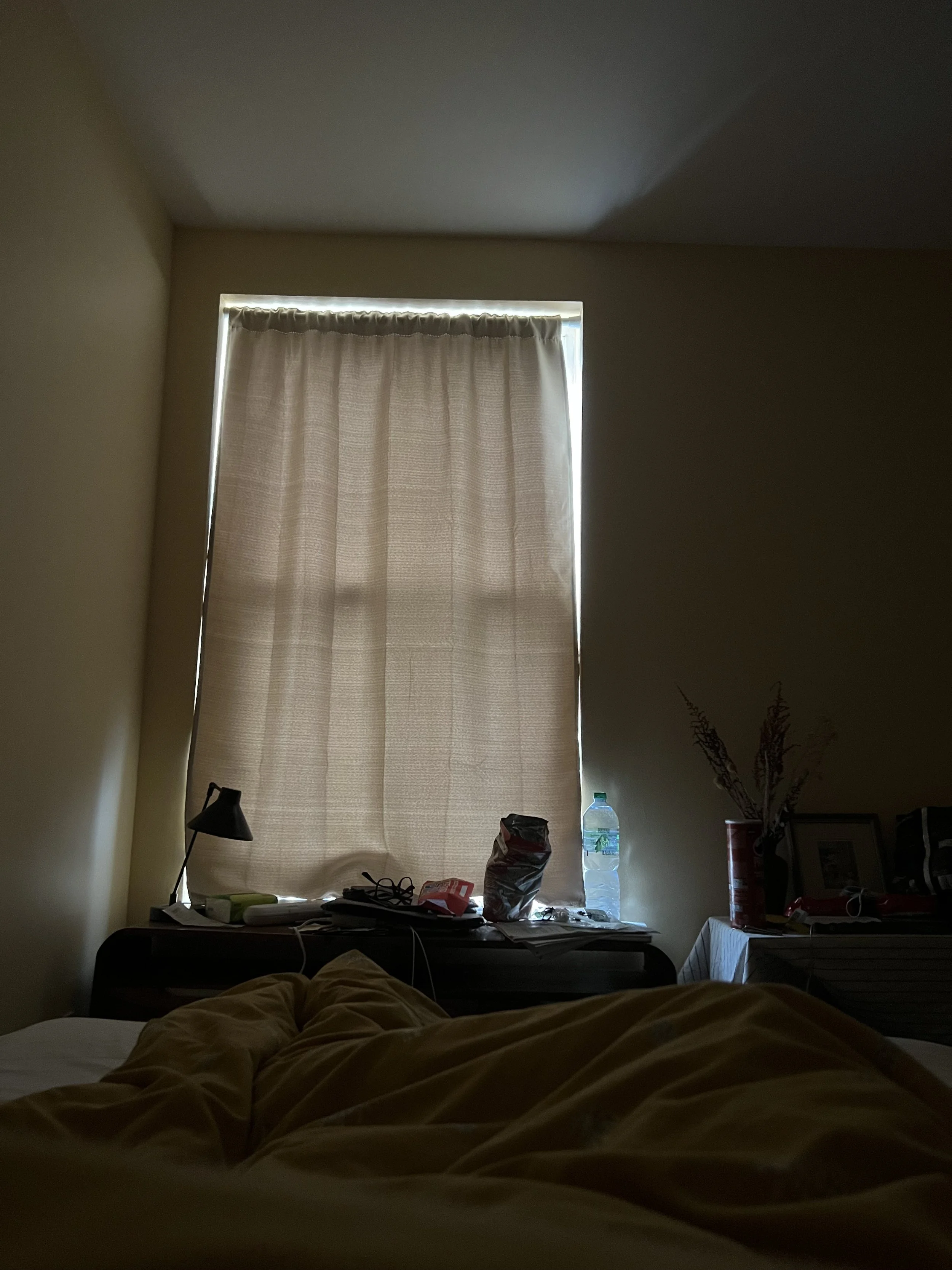 A cluttered bedroom with a bed in foreground covered with yellowish bedspread, a desk with various items and a window with closed beige curtains in the background, low lighting.