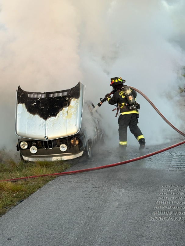 Firefighter spraying water on a car that is burning after crashing into the side of a road, with smoke rising from the vehicle.