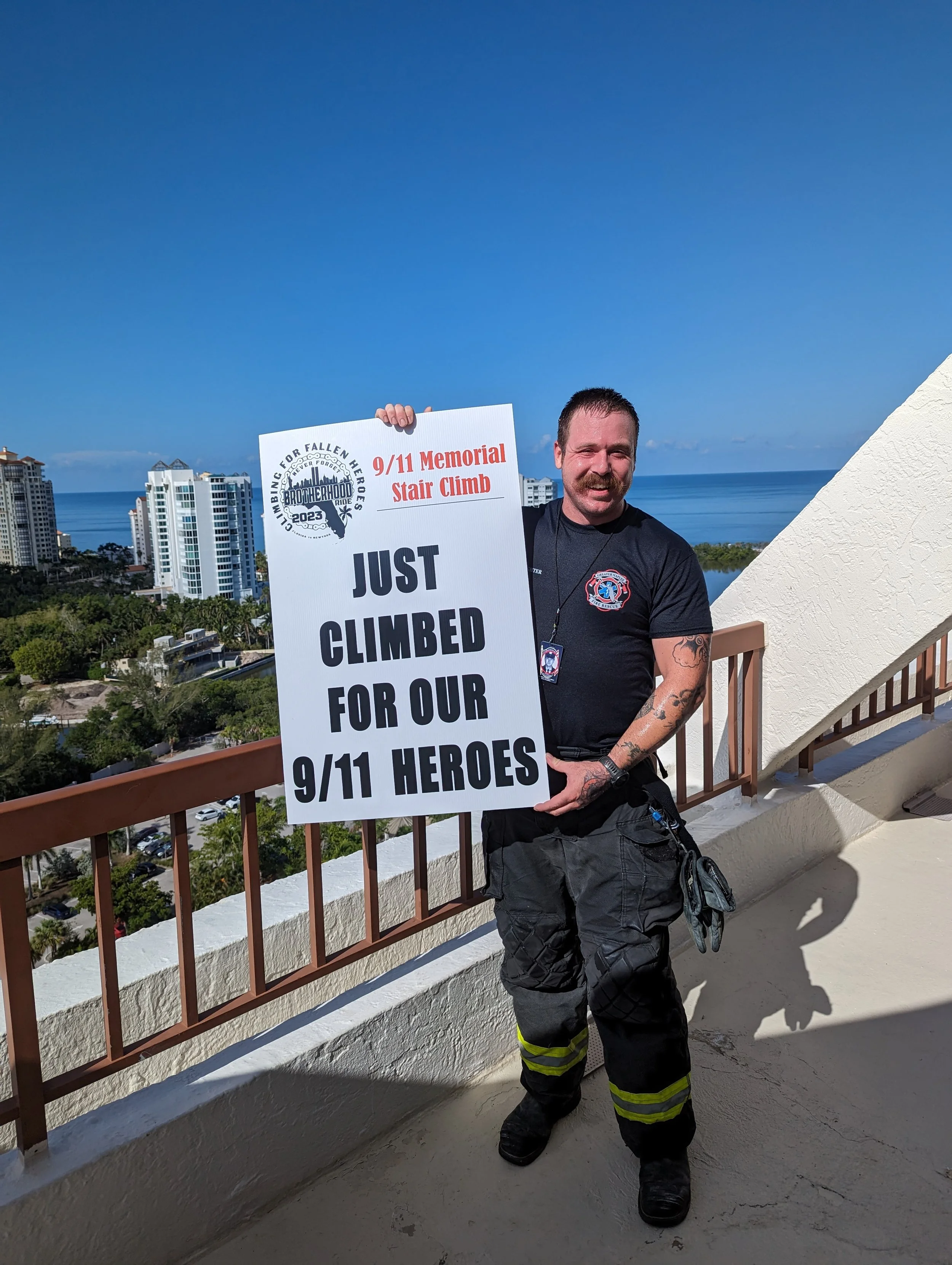 Man in firefighter gear holding a large sign on a balcony with a cityscape and ocean in the background.