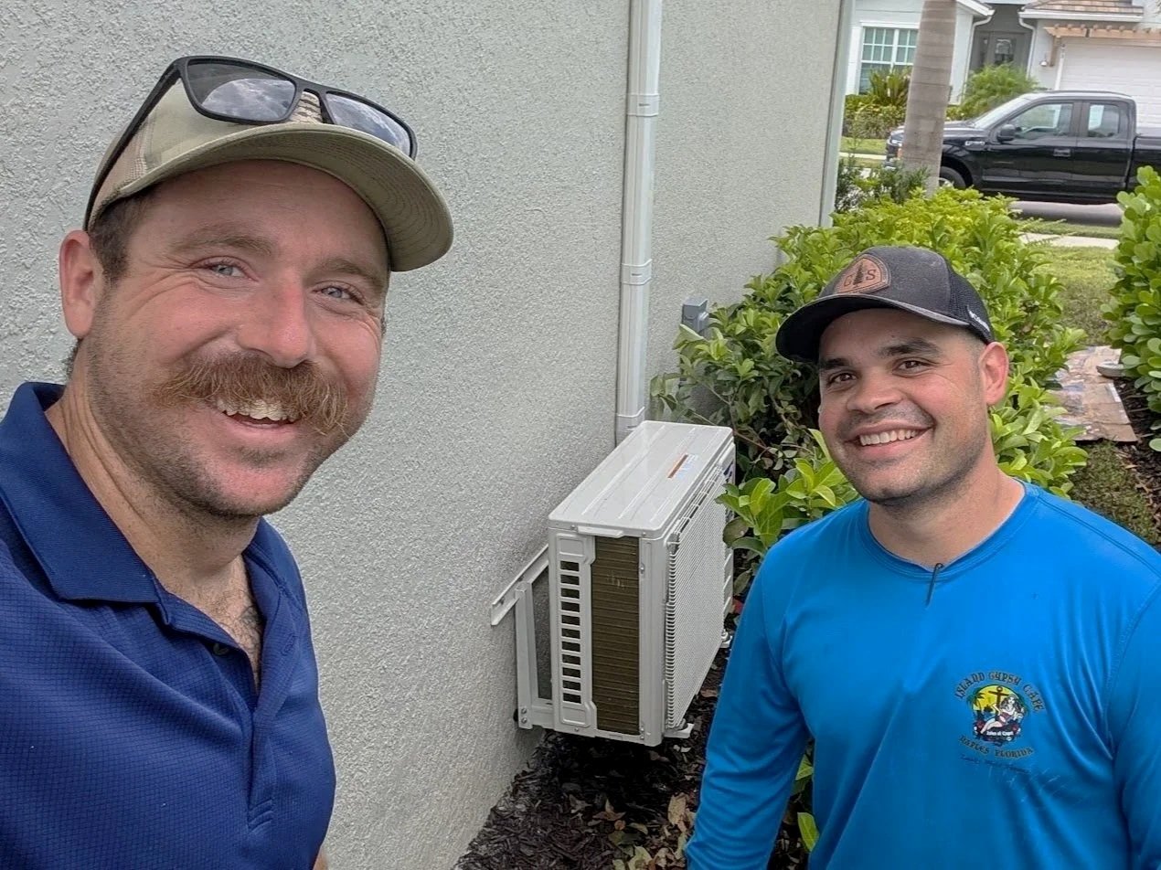 Two men smiling outdoors, standing in front of a wall with a window air conditioning unit, bushes, and trees in the background.