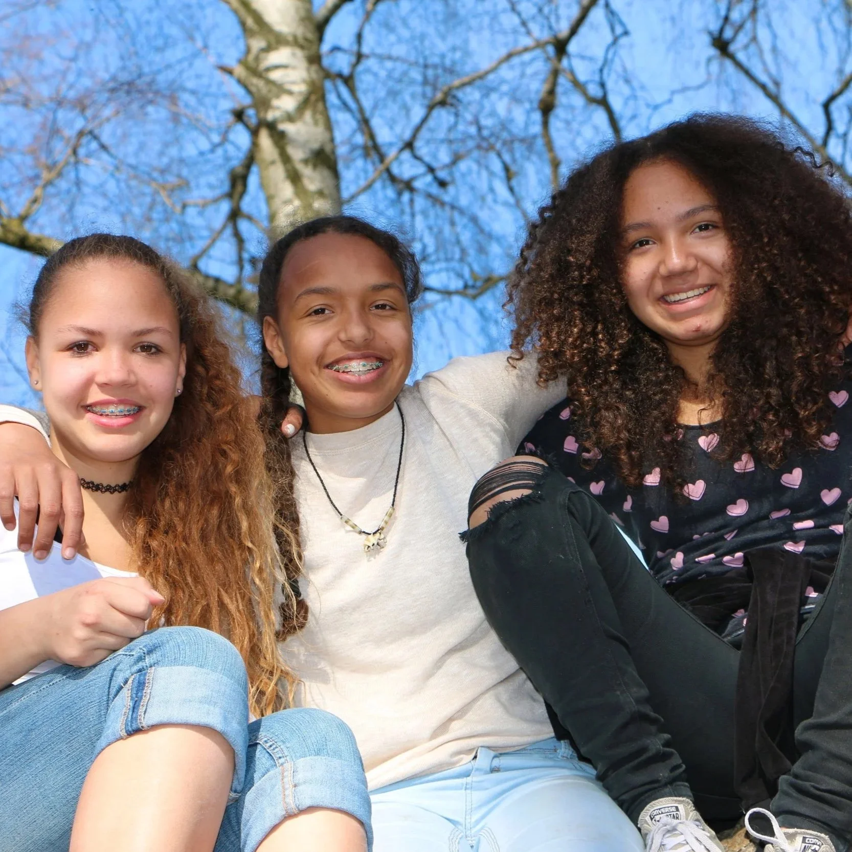 Three girls sitting outside, smiling, with trees and a blue sky in the background, dressed casually.