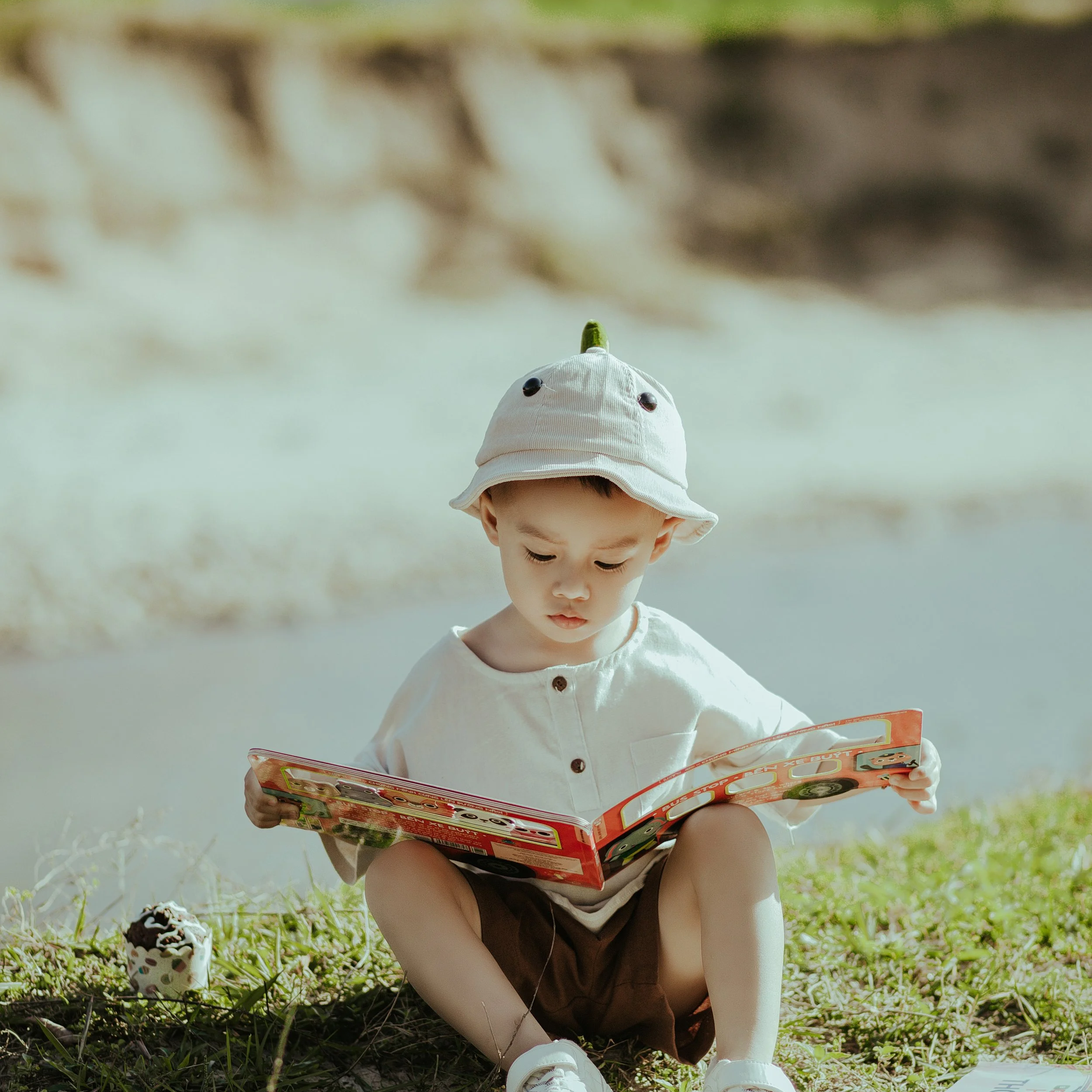 A young boy sitting on grass outdoors, wearing a white hat, white shirt, and brown shorts, reading a colorful book with pictures.