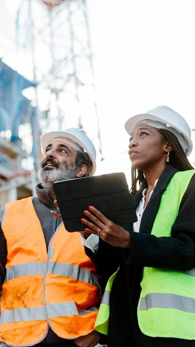 Two builders or two engineers wearing safety vests and hardhats reviewing project on a tablet at a jobsite