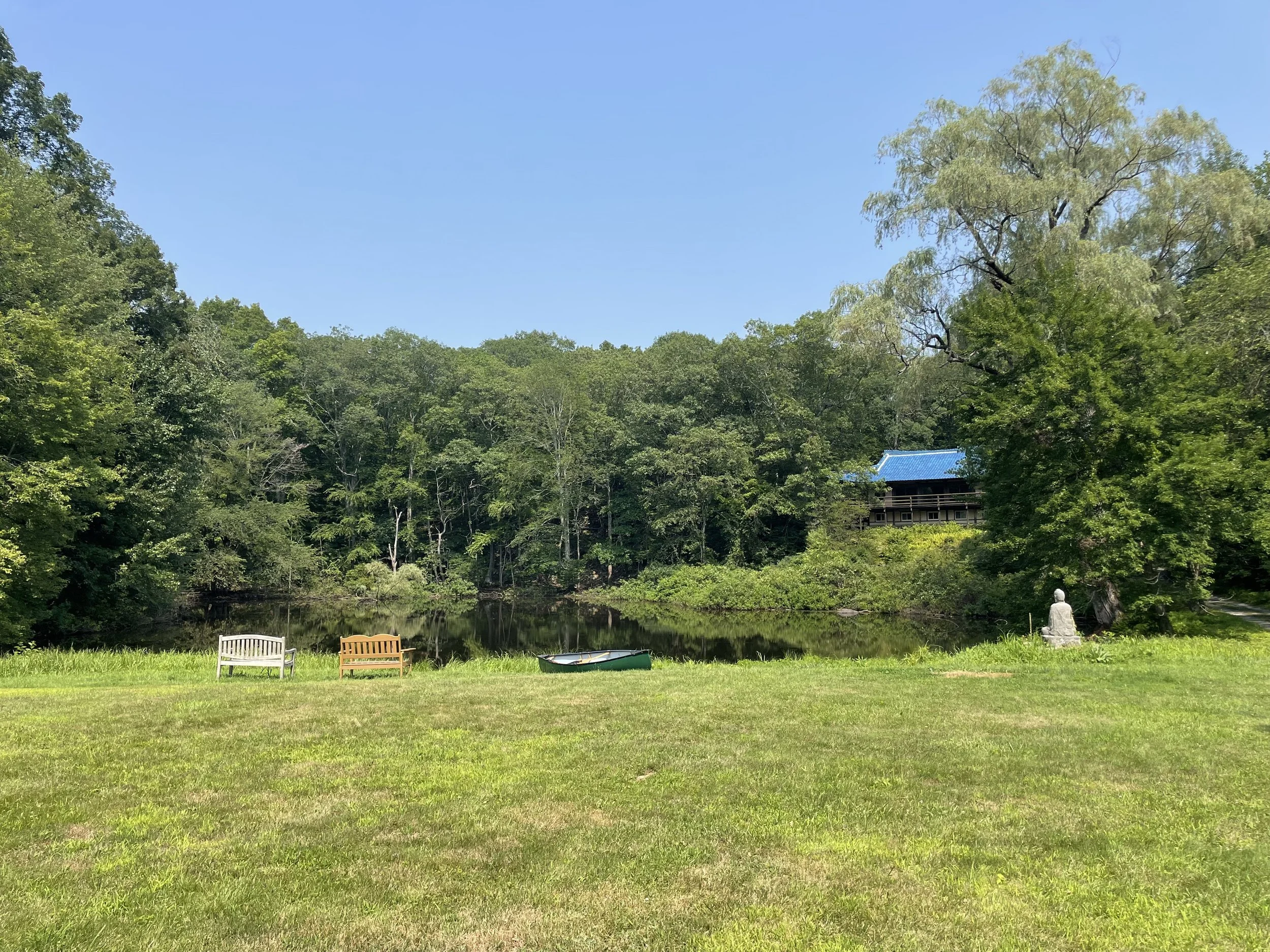 A serene outdoor scene featuring a grassy area with three empty chairs, a small boat, a statue of a person, and a tranquil pond surrounded by lush green trees under a clear blue sky.