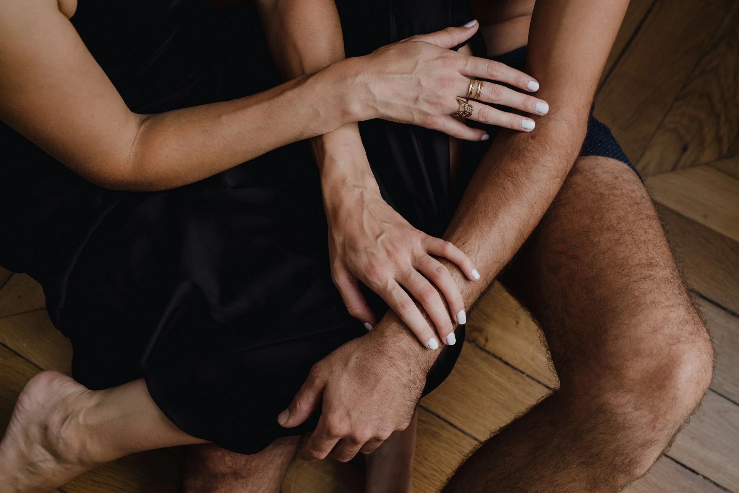 Close-up of multiple hands and legs intertwined on a wooden floor, with some hands wearing rings and painted nails.