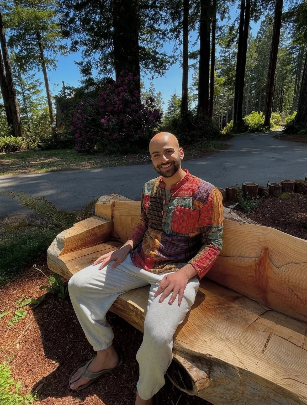A man with a beard and a bald head smiling, sitting on a carved wooden bench in a forested area with trees, plants, and a dirt path visible in the background, wearing a colorful patchwork shirt and white pants.