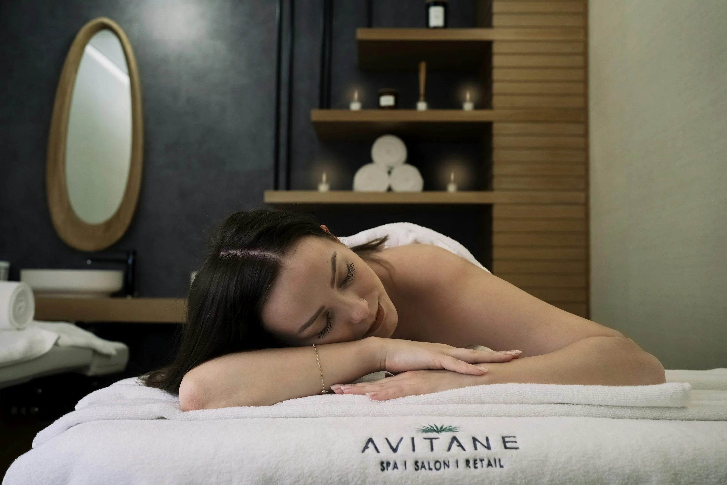 A woman with dark hair is lying face down on a massage table with her eyes closed. The room features dark and wooden walls with decorative candles and a mirror. The massage table has a towel with the words 'AVITANE' and 'Spa | Salon | Retail' on it.