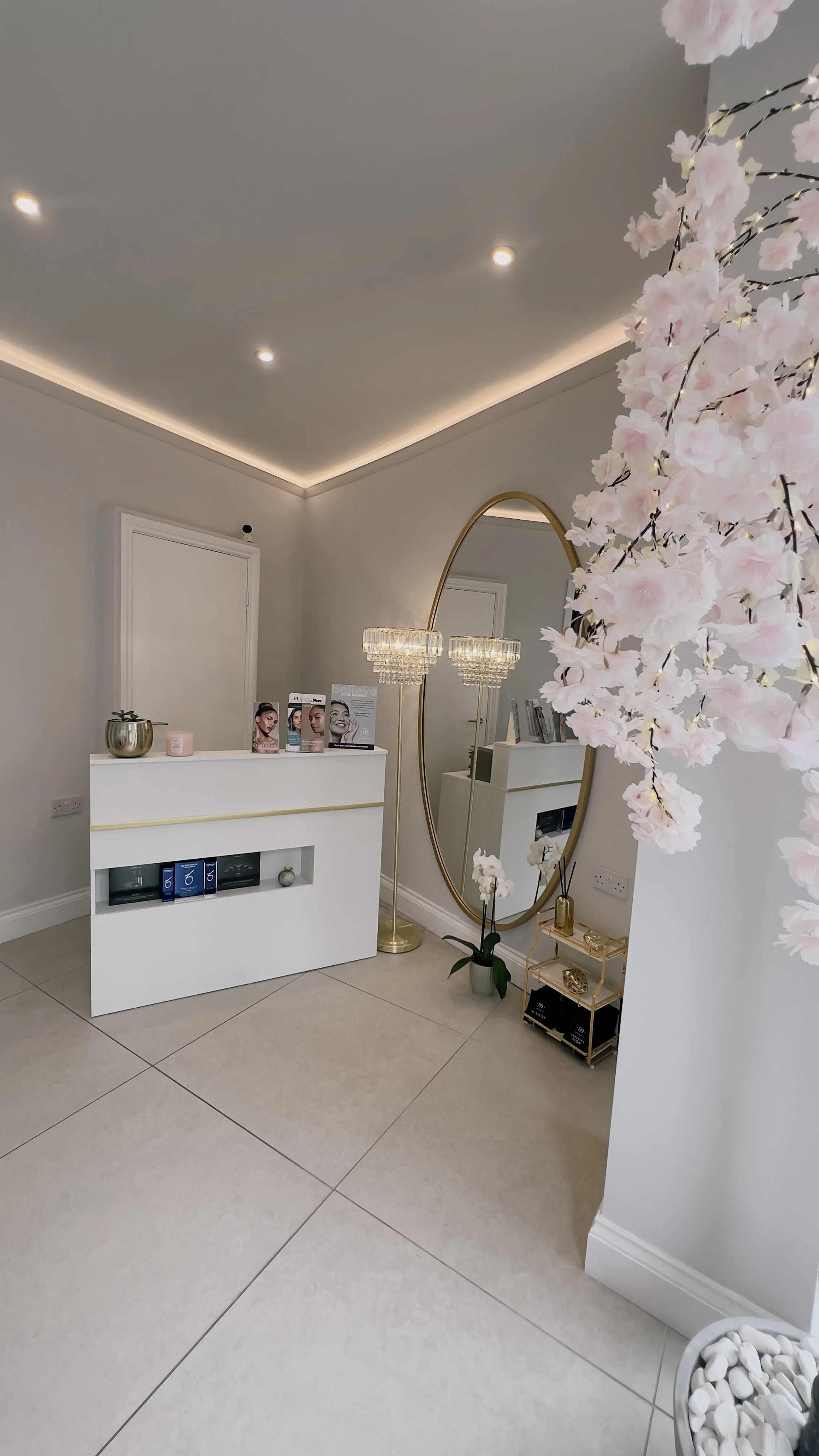 A modern, minimalistic interior space featuring white walls, large tiled floor, a white console table with decor items, a large oval mirror with a gold frame, a tall gold floor lamp with a crystal chandelier shade, and pink cherry blossom flowers in the foreground.
