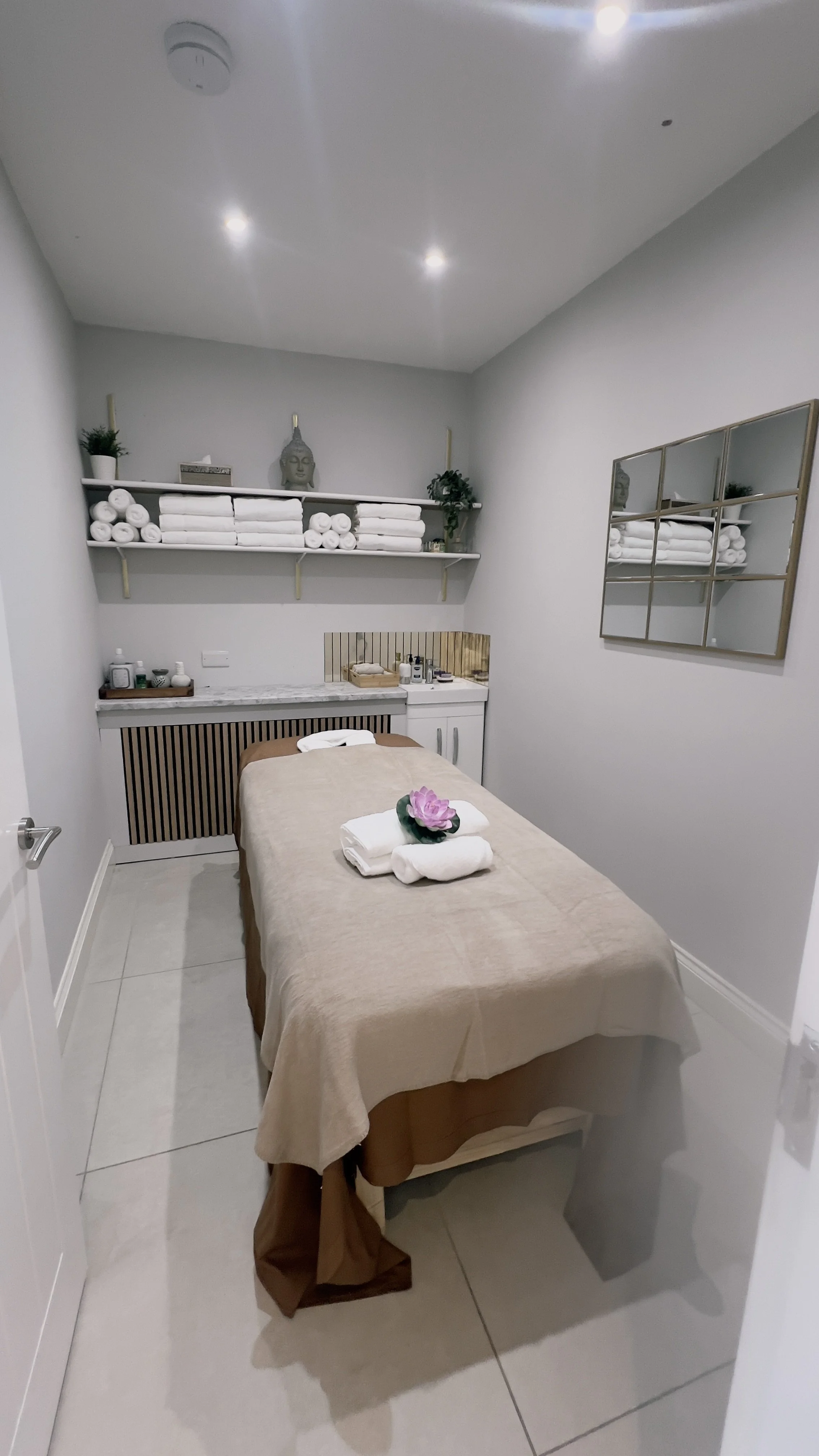 A massage table covered with a beige sheet, with folded towels and a pink flower on top, in a spa room with white walls, shelves with rolled towels, plants, and decorative items.