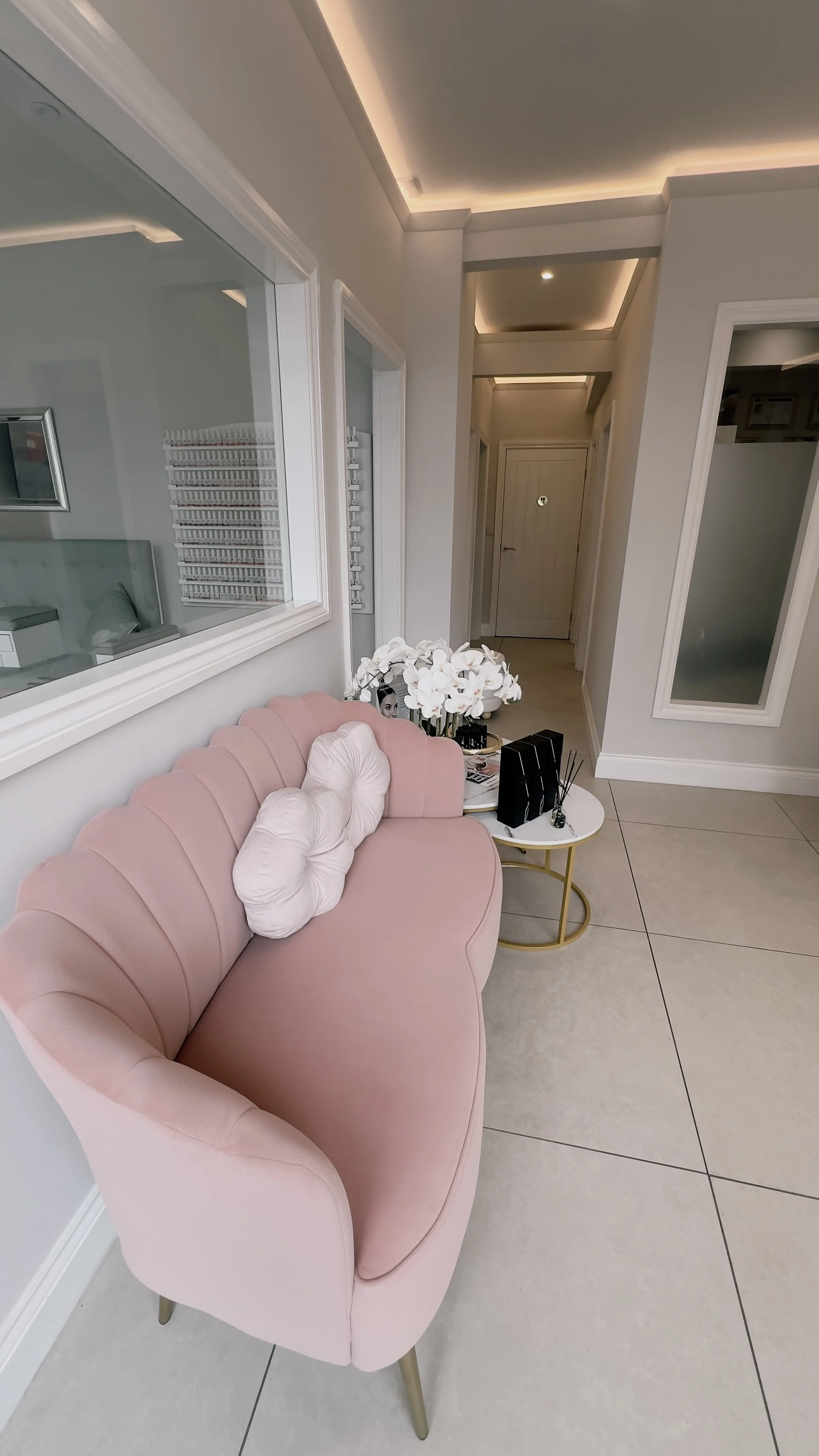 A pink velvet sofa with white tufted pillows in a modern, well-lit waiting area. A small round table next to the sofa holds a bouquet of white orchids, magazines, and black decorative items. The room has white walls, large glass windows, and a hallway with a white door at the end.
