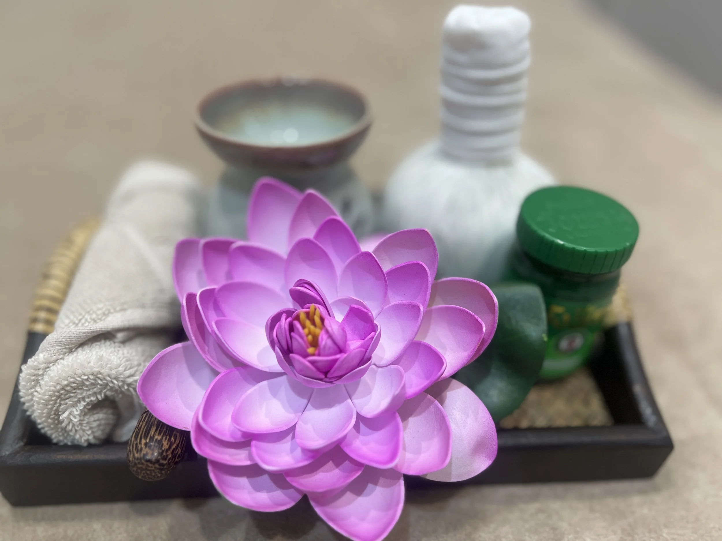 A pink artificial flower, a small ceramic bowl, a bottle with a green cap, a white towel, and a bobbin, placed on a black tray.