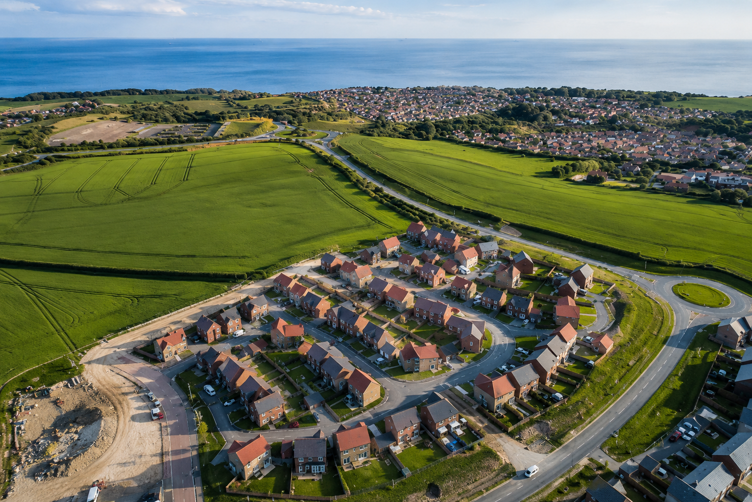 Aerial view of a coastal town with green farmland, residential neighborhoods, winding roads, and the blue sea beyond