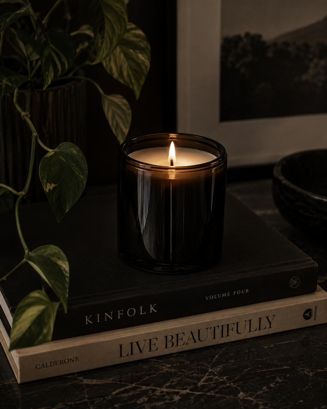 A lit candle in a dark glass holder on top of two stacked books titled 'Kinfolk' and 'Live Beautifully', with a leafy plant and a framed picture in the background.