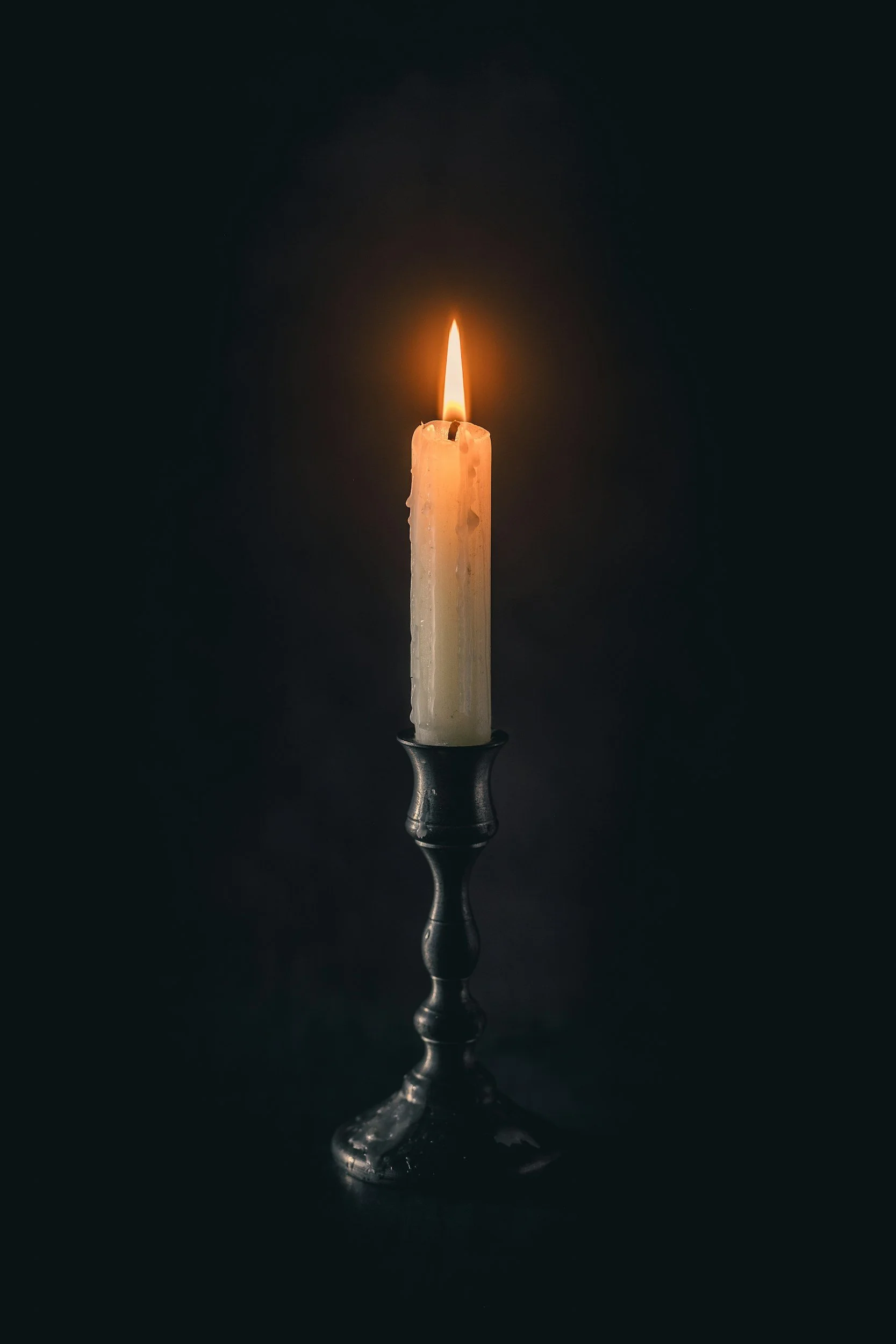 Tall white candle burning with a small flame on a black candle holder, isolated on black background.