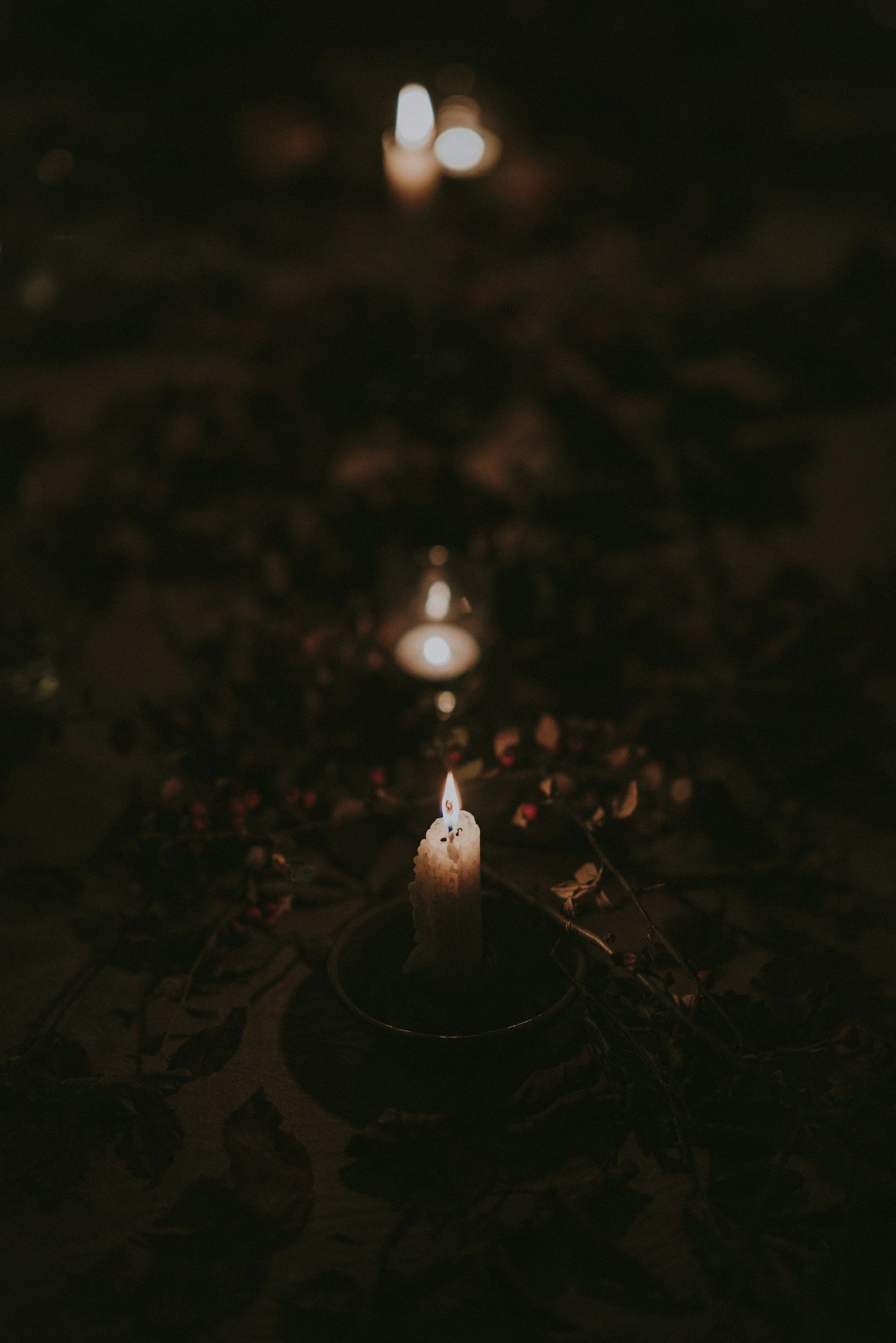 A single candle burning on a dark surface in a circular holder, with faint light reflections and surrounding dried leaves or petals.