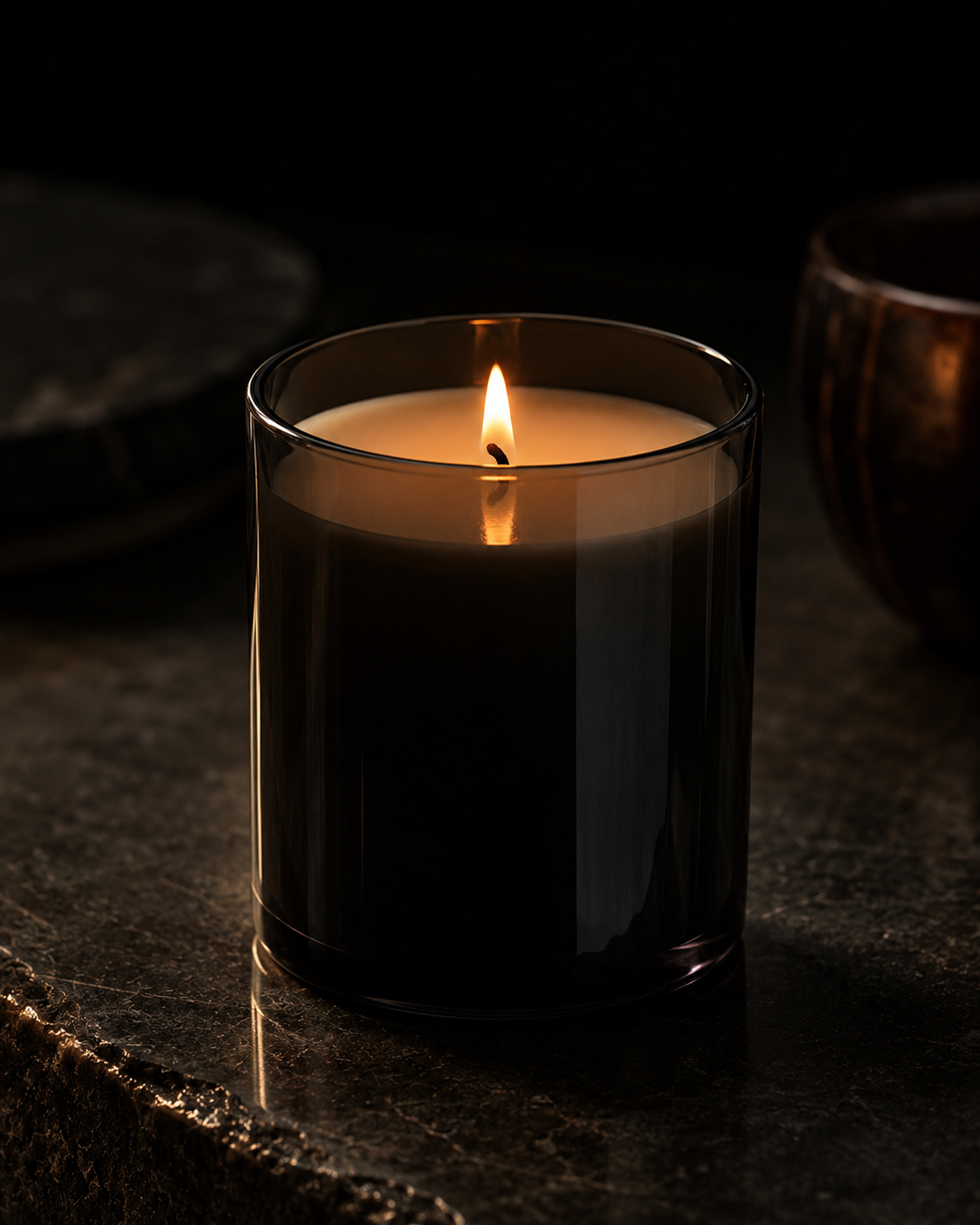 A lit candle in a glass container on a dark surface with a blurred stone and bowl in the background.
