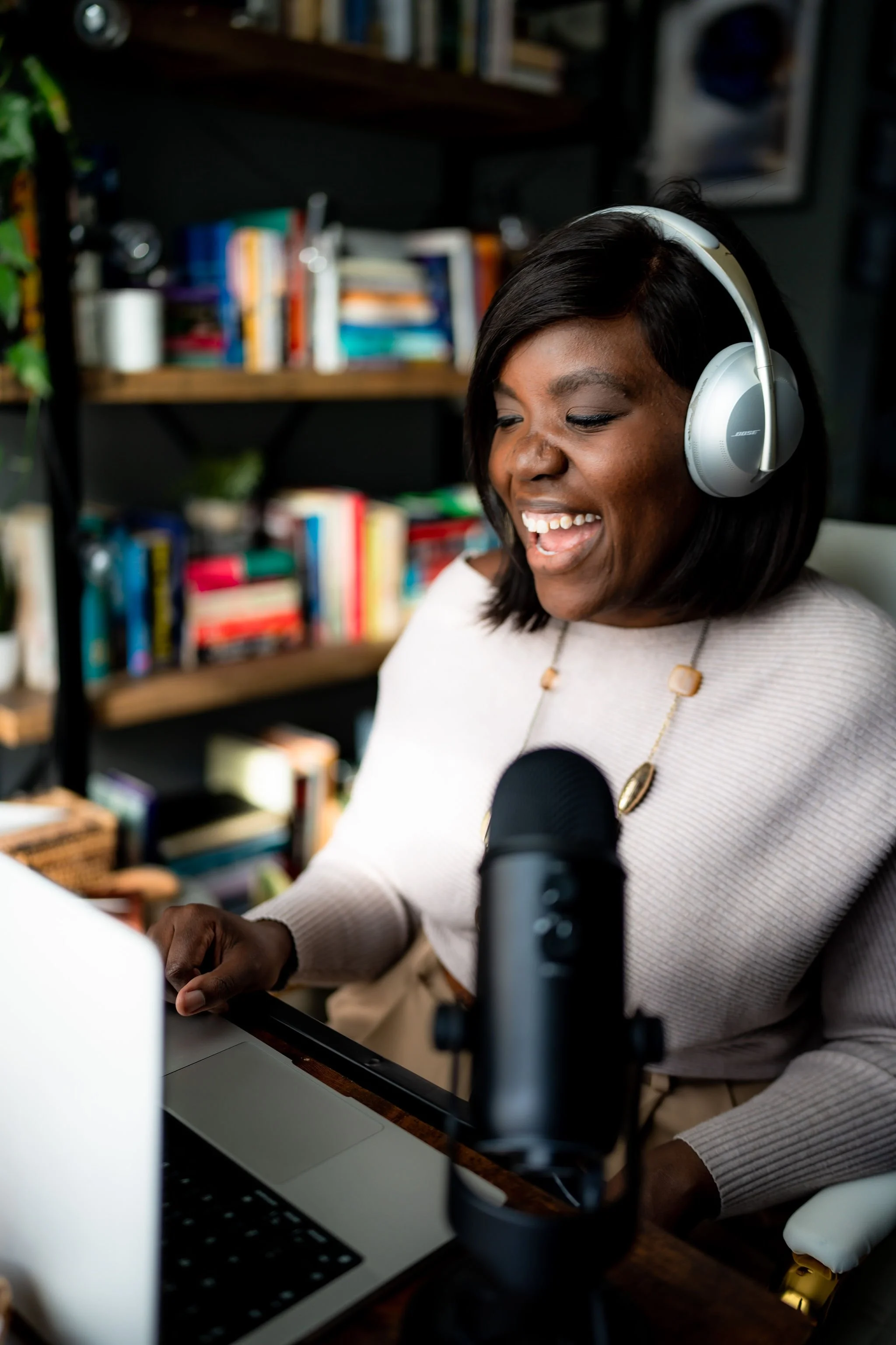A woman with dark hair wearing large Bose headphones, sitting at a desk with a microphone, a laptop, and bookshelves in the background, smiling while working in a home office.