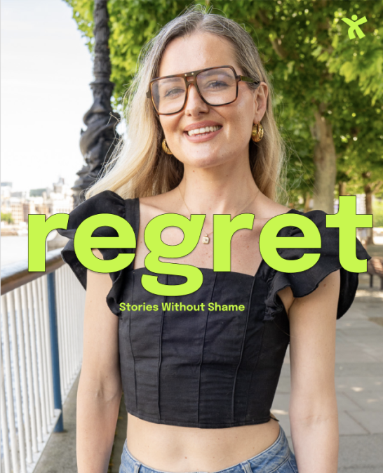 A young woman with blonde hair, wearing glasses, gold earrings, and a black crop top, smiling outdoors with green trees and a railing in the background, overlaid with the words 'regret' and 'Stories Without Shame' in bright green text.