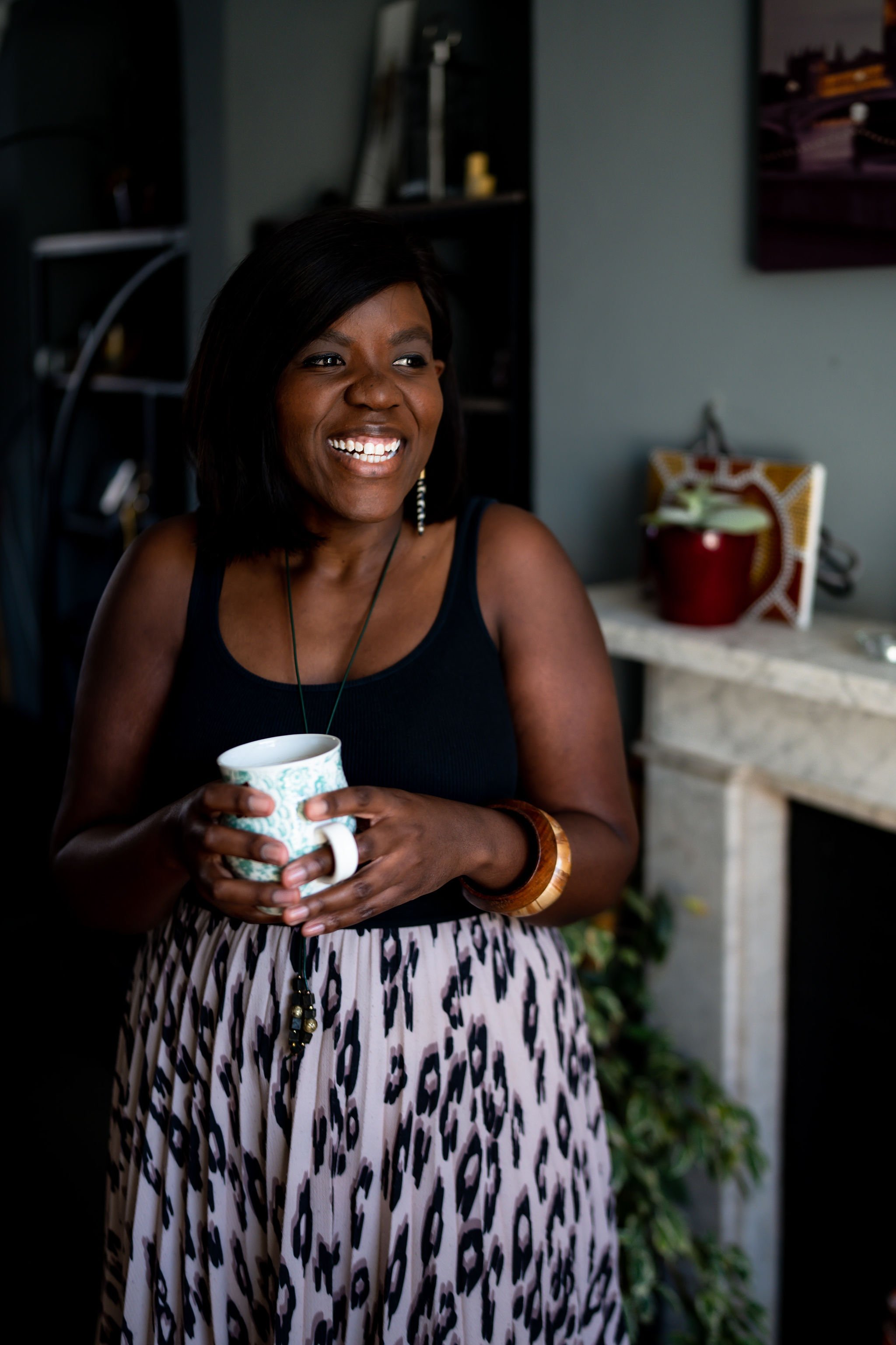 A woman with dark hair smiling, holding a white and teal patterned mug, wearing a black top, a patterned skirt, and jewelry, standing in a room with shelves, plants, and artwork.