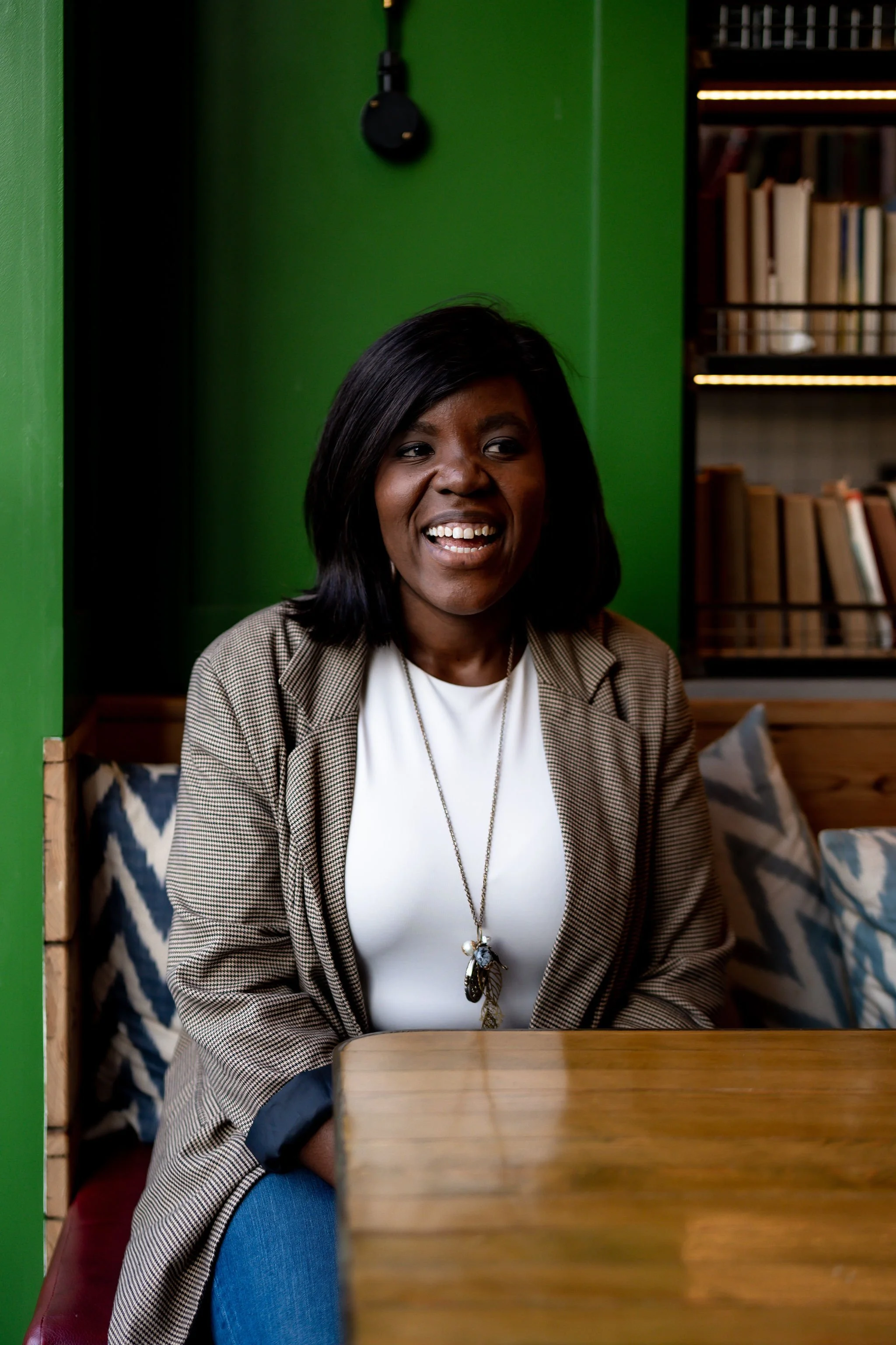 A woman with dark hair smiling, wearing a beige checkered blazer over a white shirt, sitting at a wooden table in a room with green wall and bookshelf in the background.