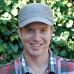 A young man wearing a gray cap and plaid shirt, smiling outdoors with greenery in the background.