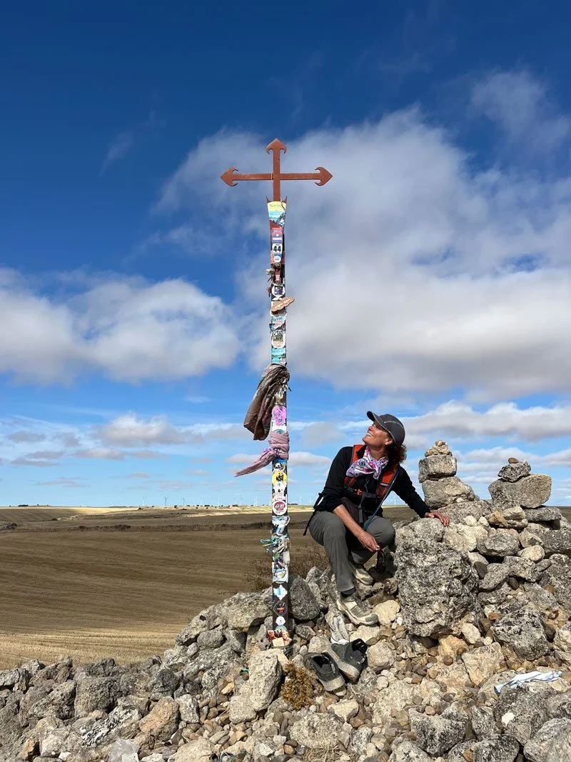 A woman sitting on a pile of rocks next to a tall, decorated cross with a blue sky and clouds in the background.