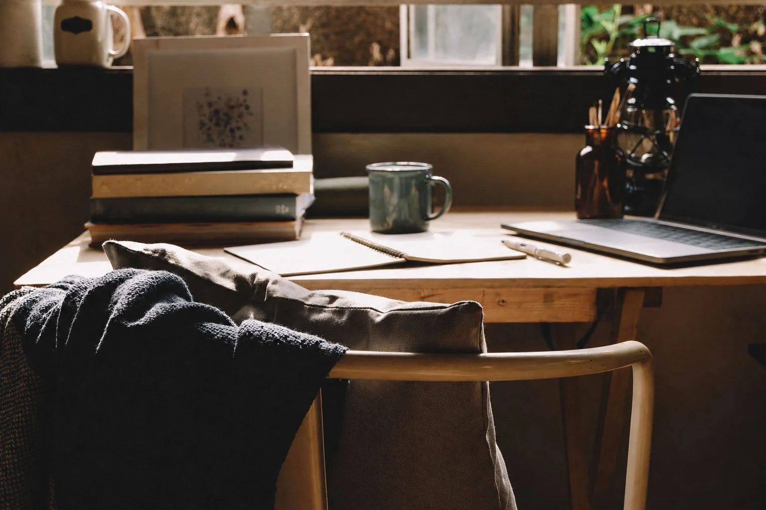 A cozy wooden desk illuminated by natural sunlight, with a laptop, a notebook, a pen, a mug, and a stack of books, situated in front of a window overlooking greenery.
