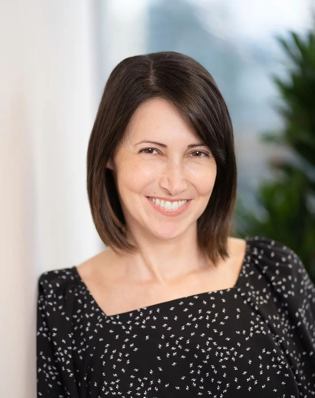 A woman with shoulder-length dark brown hair smiling, wearing a black dress with white floral pattern, in an indoor setting with blurred greenery and light background.
