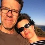 A man and woman taking a selfie outdoors with a clear blue sky and ocean in the background.