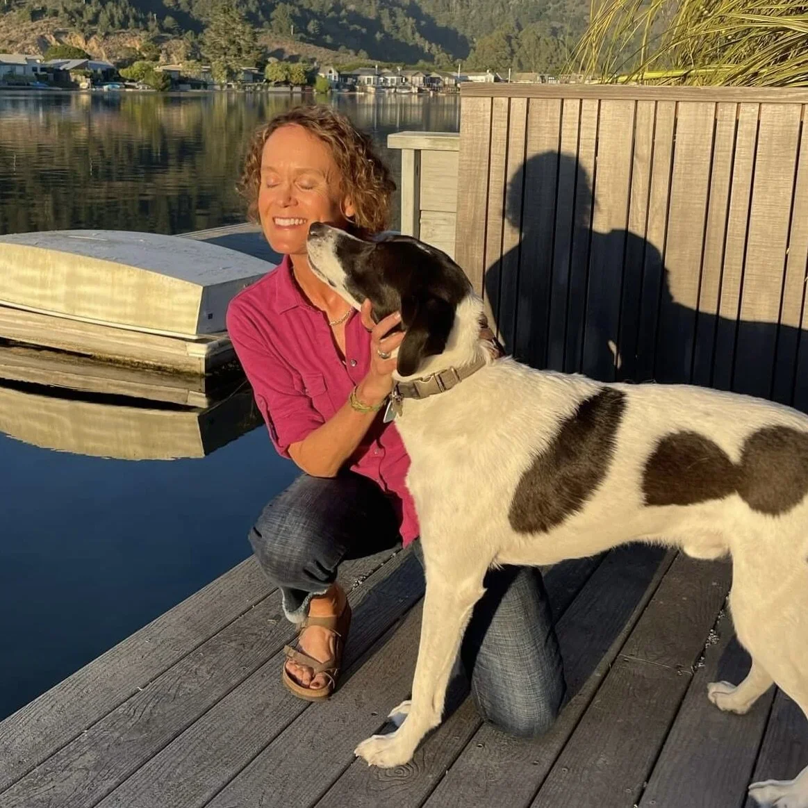 A woman with curly hair smiling with closed eyes, in a pink shirt, kneeling on a wooden dock, kissing a large black and white dog near a calm lake during sunset.