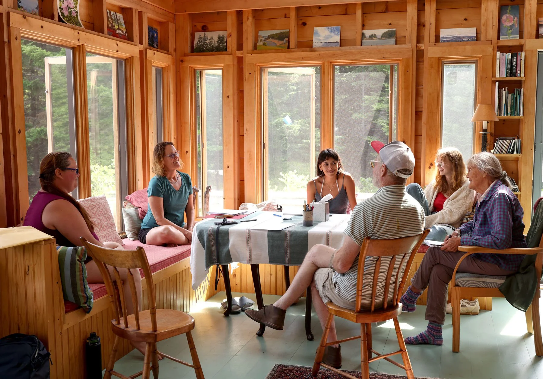 A group of seven people gathered around a table in a sunlit wooden room, engaging in conversation. The room has large windows with a view of trees outside, wooden walls decorated with paintings, and a bookshelf.
