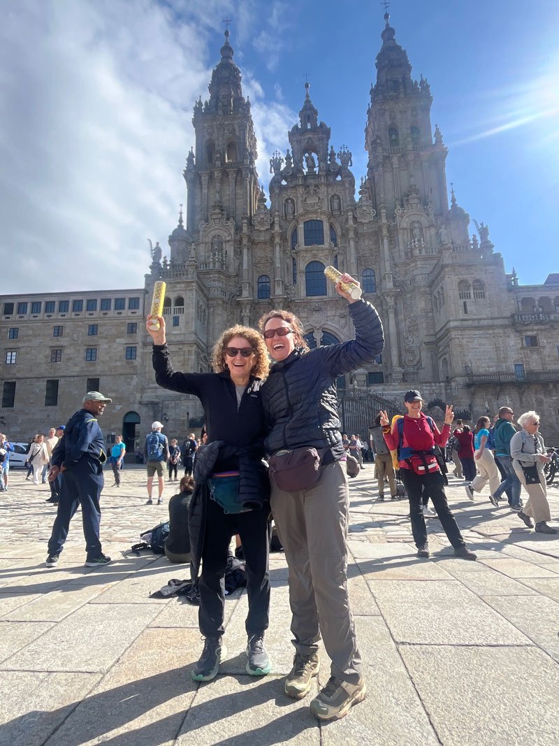 Two women smiling and holding ice cream cones in front of a large historic cathedral, with a crowd of tourists around them.