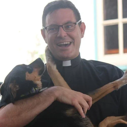 A man wearing glasses and a clergy shirt holding a black and brown dog with one paw on his cheek, inside a bright room with a window in the background.