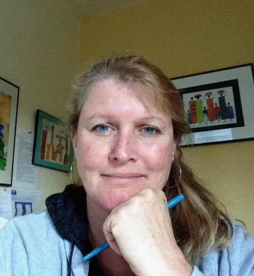 A woman with blonde hair and blue eyes sitting at a desk, holding a blue pen near her chin, in an office with colorful framed artwork on the wall.