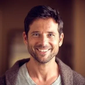 A smiling man with dark hair and beard wearing a gray shirt and brown cardigan