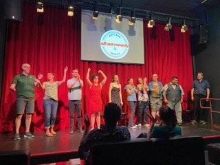 A group of people standing on stage holding hands and raising their arms together during a performance or event, with an audience seated in front and a red curtain in the background.
