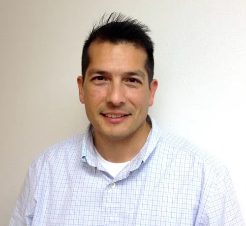 A man with short dark hair, wearing a light blue checkered button-up shirt, smiling, standing against a plain white wall.