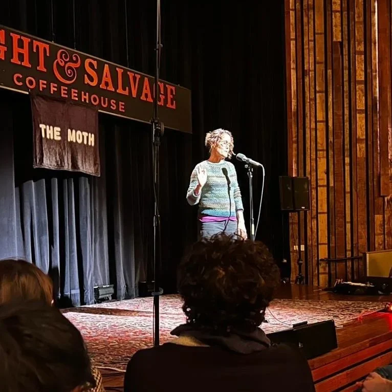 A woman with curly gray hair standing on a stage, holding a microphone, and performing in a comedy club. The stage has a backdrop with a sign that reads 'Night & Salvage Coffeehouse' and a curtain with a banner that says 'The Moth.' Audience members are visible in the foreground.