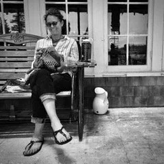 A woman sitting on a bench outdoors, reading a book, wearing sunglasses, a patterned shirt, and sandals.