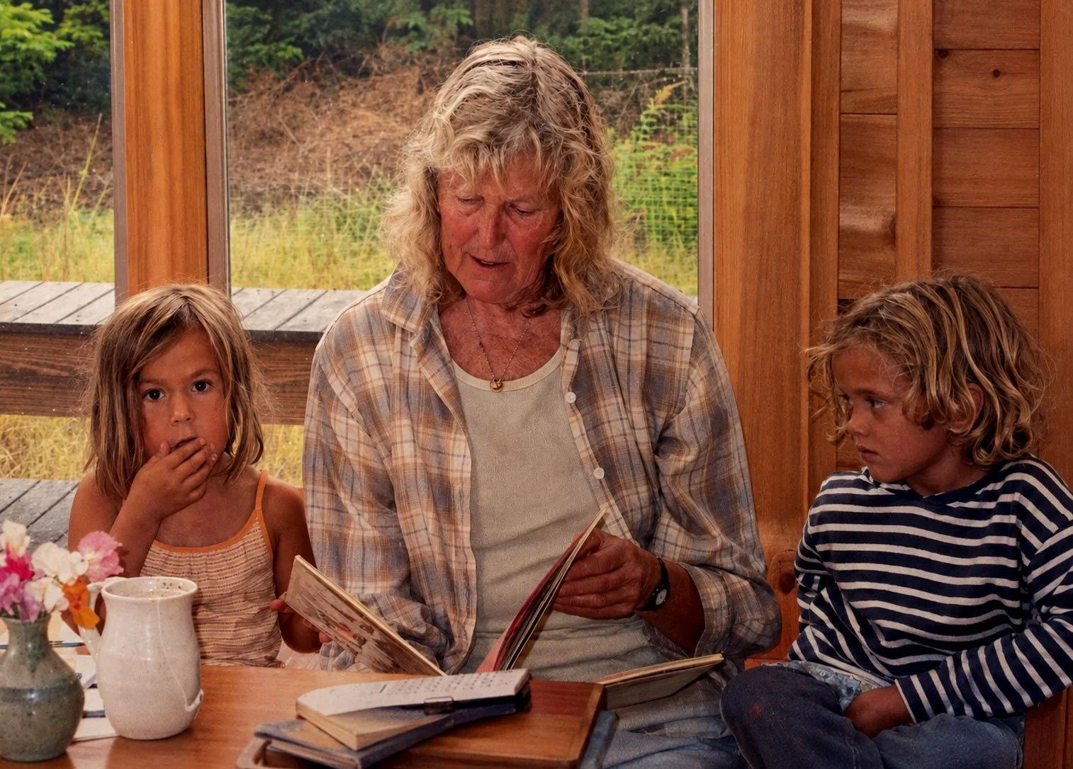 An elderly woman reading a book to two young children, a girl and a boy, sitting at a wooden table inside a room with a window showing the outdoors.