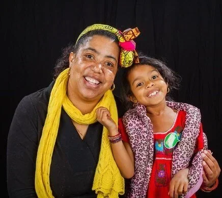 A woman and a young girl smiling and posing together against a black curtain background. The woman is wearing a yellow headscarf and a black shirt, while the girl is wearing a red dress with a patterned jacket.