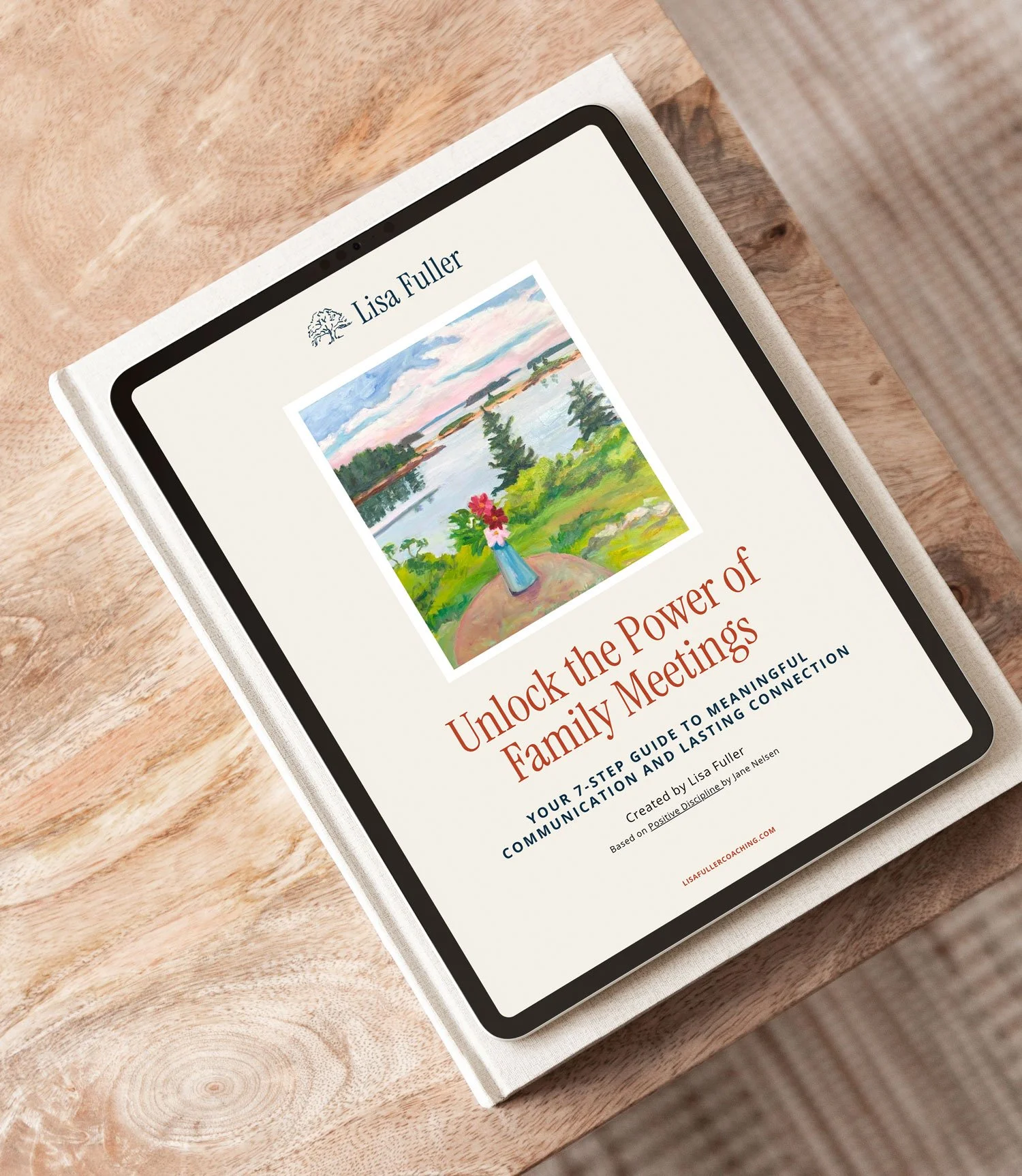 Digital tablet displaying a book cover titled "Unlock the Power of Family Meetings" by Lisa Fuller, with a landscape painting of a lake and trees, placed on a wooden table.