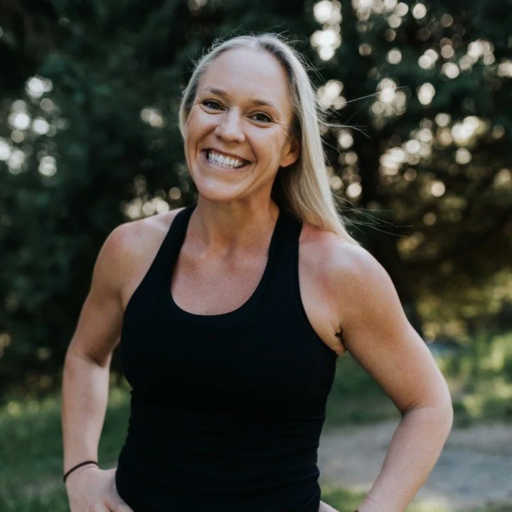A smiling woman with blonde hair wearing a black sleeveless top outdoors in a natural setting.