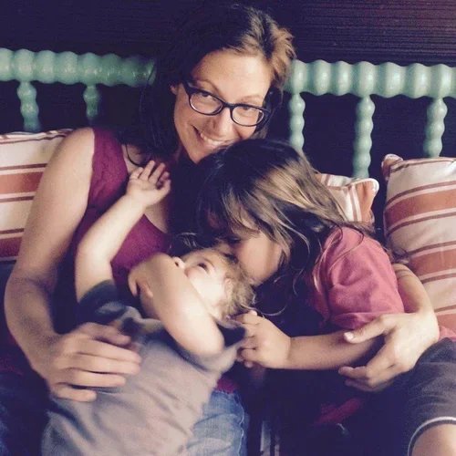 A woman with glasses and two children, one girl and one boy, smiling on a bed with striped and solid pillows, sharing a joyful moment.