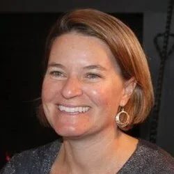 A smiling woman with short, reddish-brown hair, wearing earrings and a dark top.