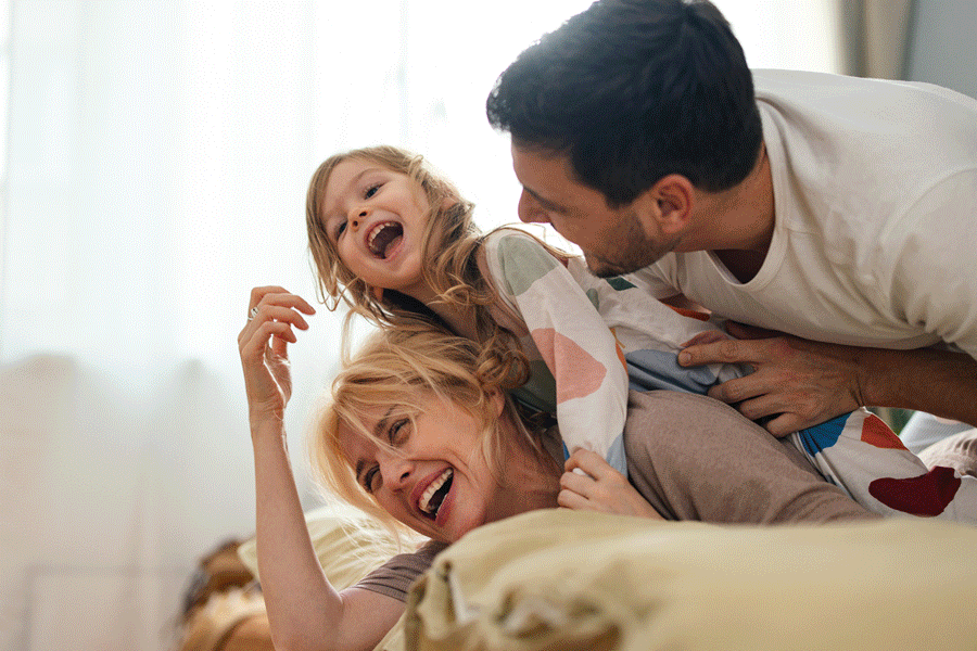 A family of three playing and laughing together on a bed in a bright room.