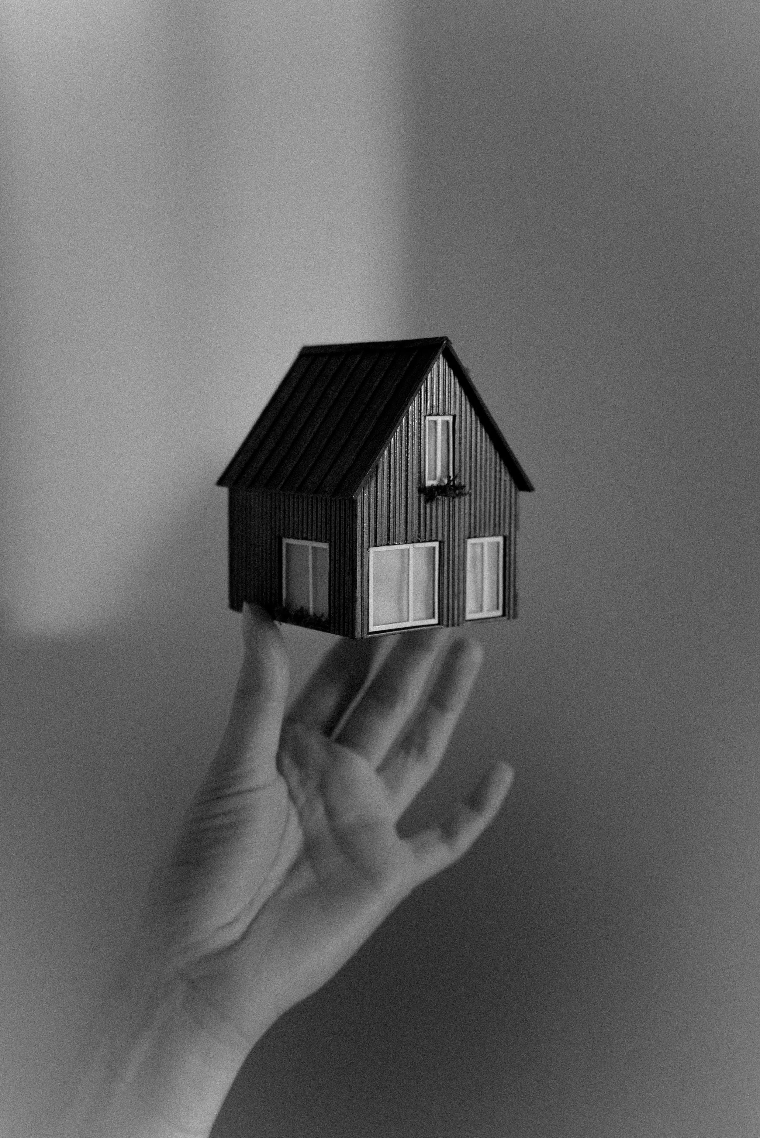 A black-and-white photo of a hand holding a small model house with a pitched roof and large windows.