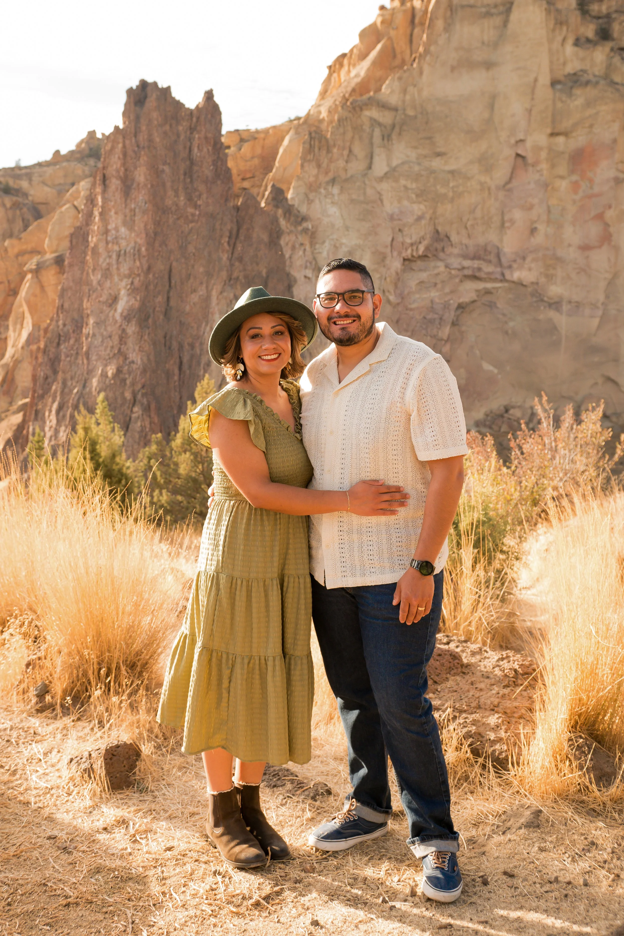 A happy couple smiling and hugging each other outdoors in a desert landscape with large rocks and yellow grass, during daytime.
