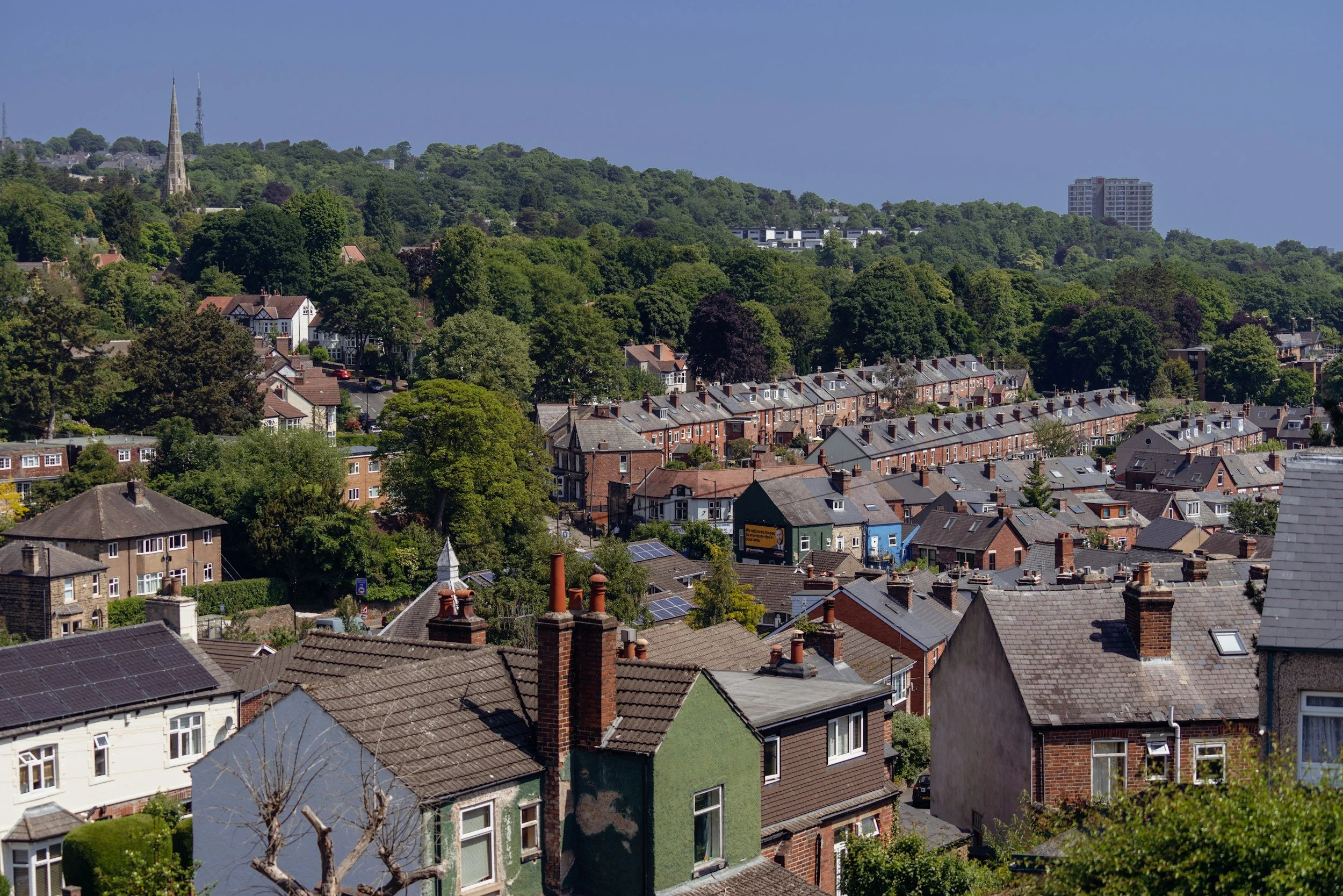 A hillside suburban neighborhood with a mix of houses and trees, some rooftops have solar panels, with larger buildings and green hills in the distance under a clear blue sky.