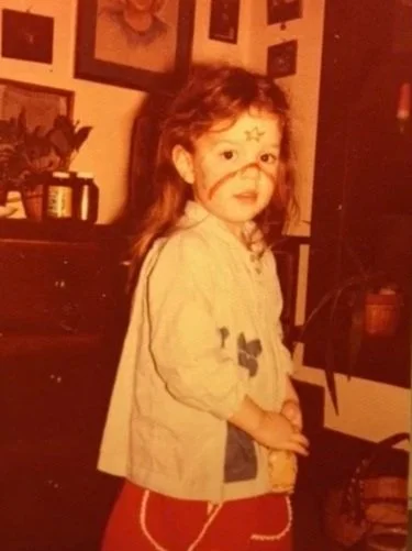A young girl with face paint of a star and butterfly, wearing a light jacket and red pants, standing indoors with framed pictures on the wall behind her.
